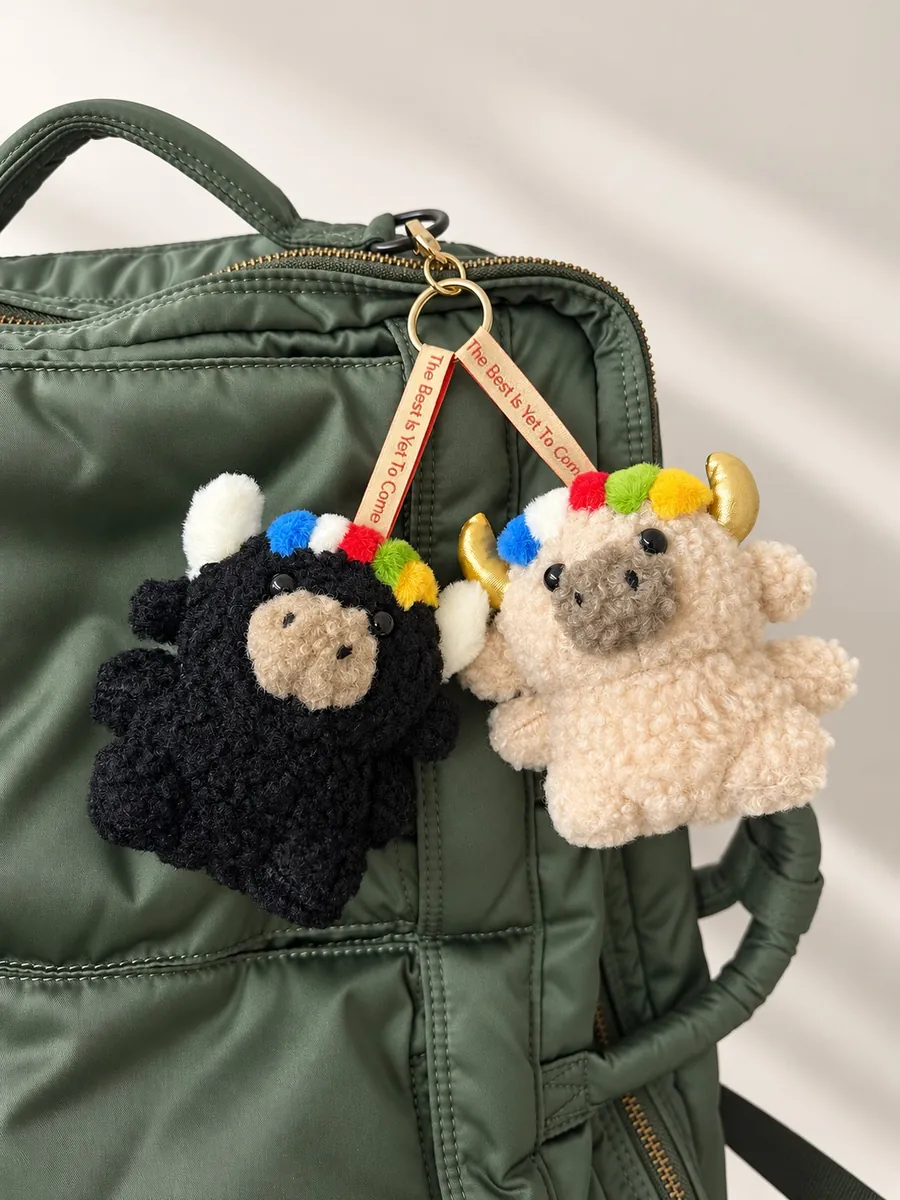 Tashi and Dele hanging side by side on a deep-green backpack, straps reading The Best Is Yet To Come — 綠色背包上並排掛著的扎西與德勒
