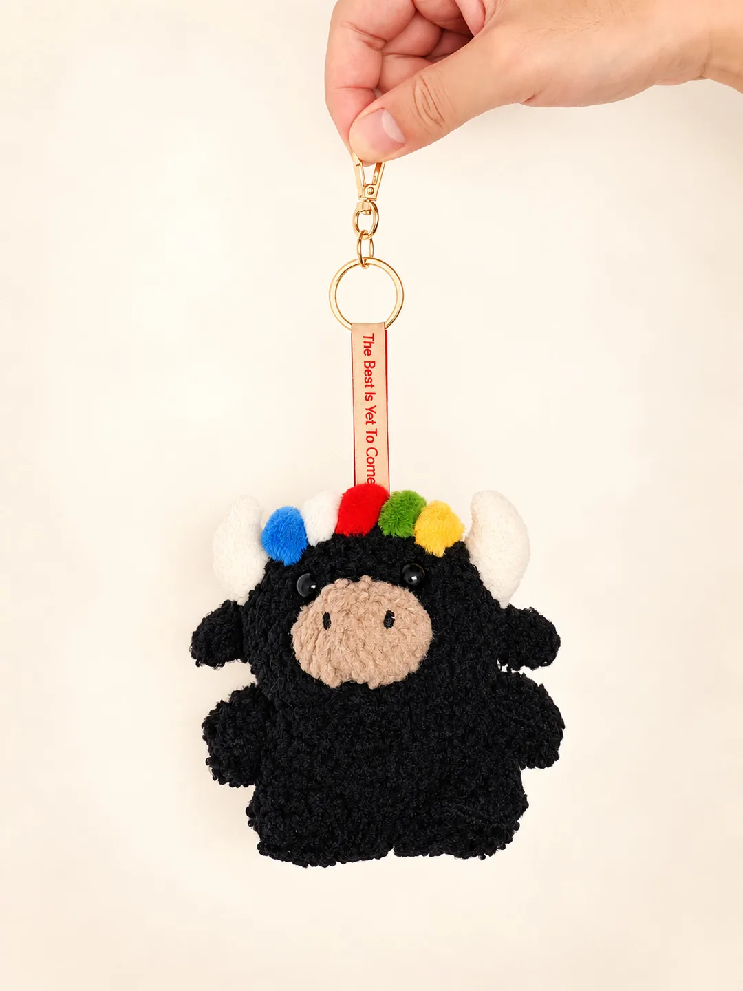 Studio portrait of Tashi the black yak plush keychain with white horns and a five-colour prayer-flag fringe, held up by hand by its gold clasp against a cream background — 黑色扎西小氂牛掛件，白角配五色經幡劉海