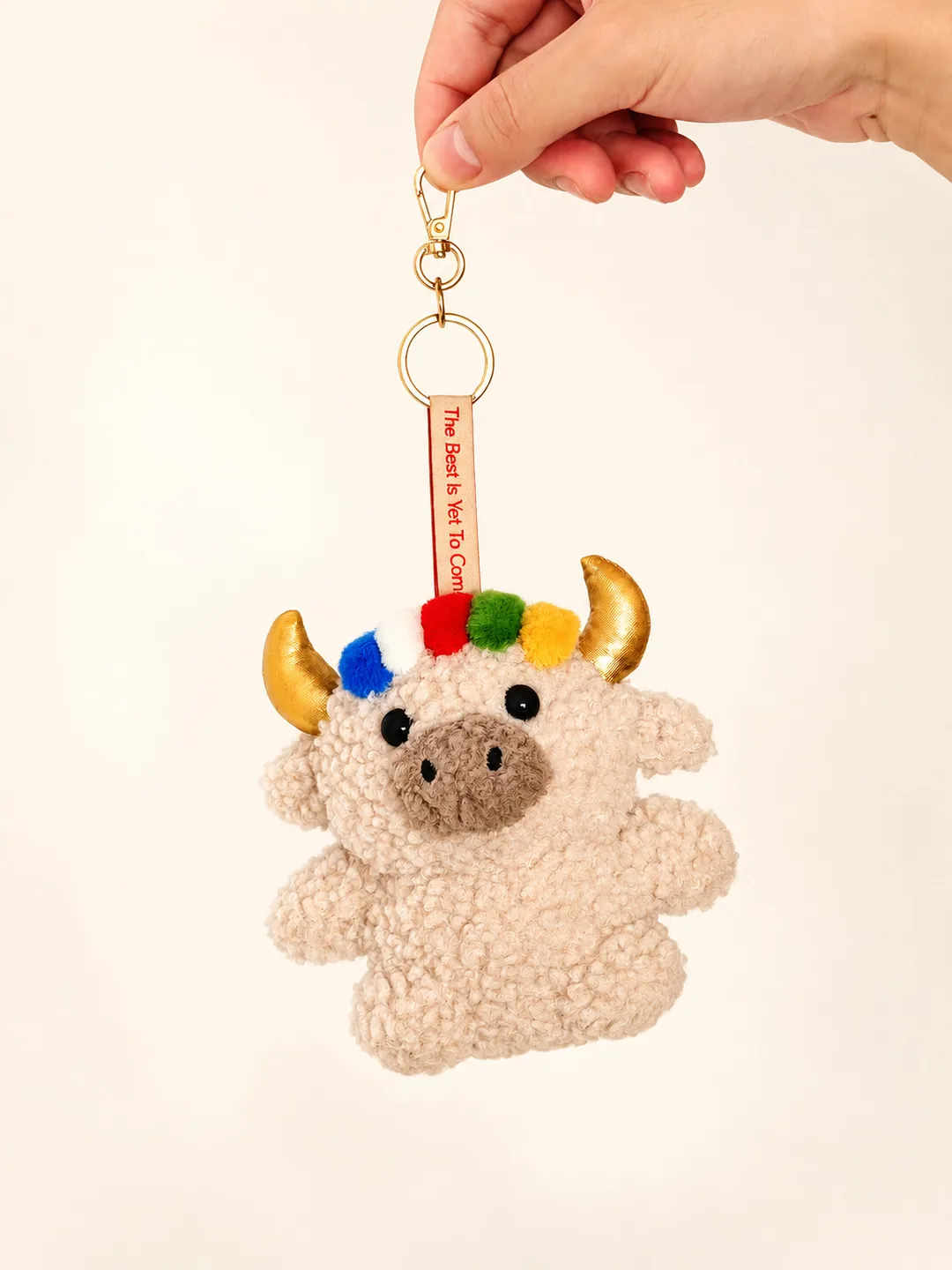 Studio portrait of Dele the cream yak plush keychain with golden horns and a five-colour prayer-flag fringe, held up by hand by its gold clasp against a cream background — 奶白色德勒富貴牛掛件，金角配五色經幡劉海